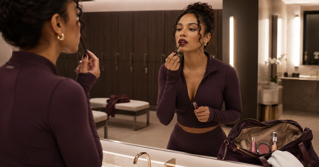 Woman in workout wear applying bold lipstick in a gym mirror with makeup products nearby